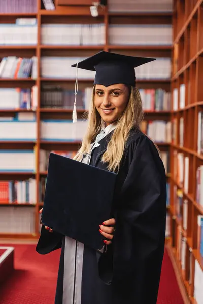 若くて笑顔の卒業生は 彼女の学士号を保持し 卒業中に図書館の中にキャップとガウンを身に着けています — ストック写真
