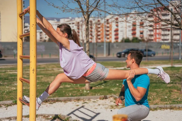 Flexible sporty girl stretching her legs. She put one leg on her male friend shoulder and the other on horizontal metal bar