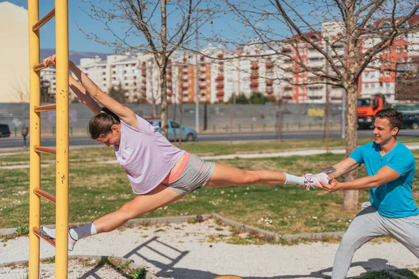 Good looking male person helping his female friend stretching her legs. He is pulling her left leg while she is holding a metal bar with her hands and the other leg