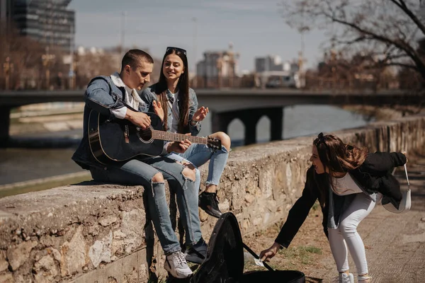 ブルネットガールは 川に座っている間にギターを演奏し歌うアーティストにお金を与えています — ストック写真