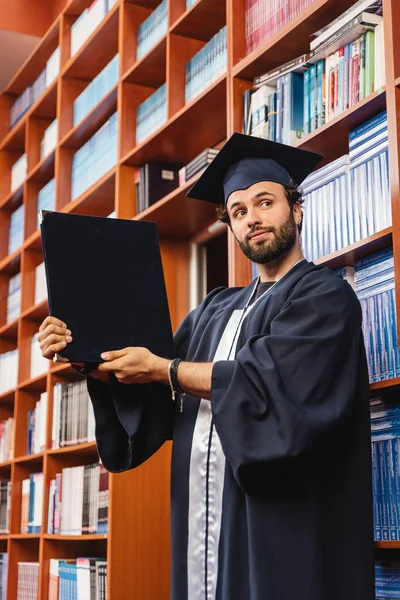 Genç Mutlu Mezun Kep Cübbe Giyiyor Lisans Diplomasını Kütüphanede Tutuyor — Stok fotoğraf