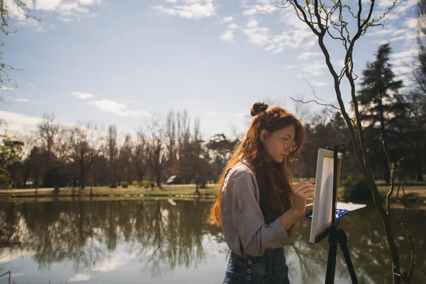 湖の近くの公園で立ってリラックスしながらキャンバスに絵を描く美しいジンジャーガール — ストック写真