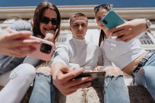 Three best friends holding their phones while sitting in front of the white building