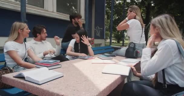 大学キャンパスの外の都市環境で知識を共有する多種多様な学生グループが オープンブックでいっぱいのベンチに座っています — ストック動画