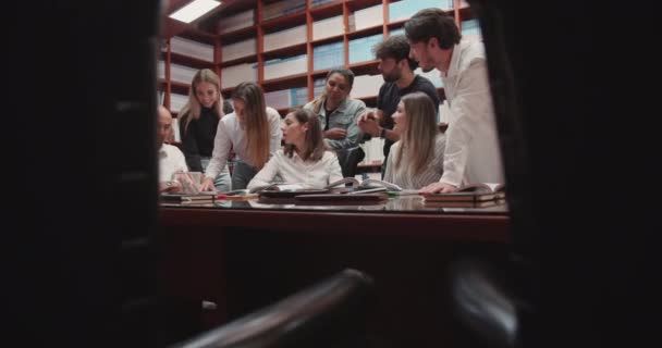 在一个忙得不可开交的大学图书馆里 一群各种各样的大学生围坐在一张桌子旁 认真地和他们的教授商量着 — 图库视频影像