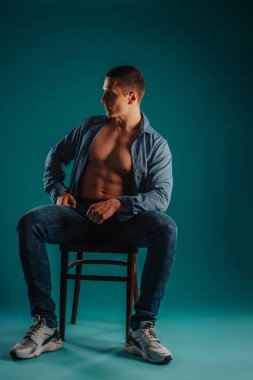 Fit shirtless guy with unbuttoned shirt sitting on a chair posing in studio on a turquoise background. He is looking aside. clipart