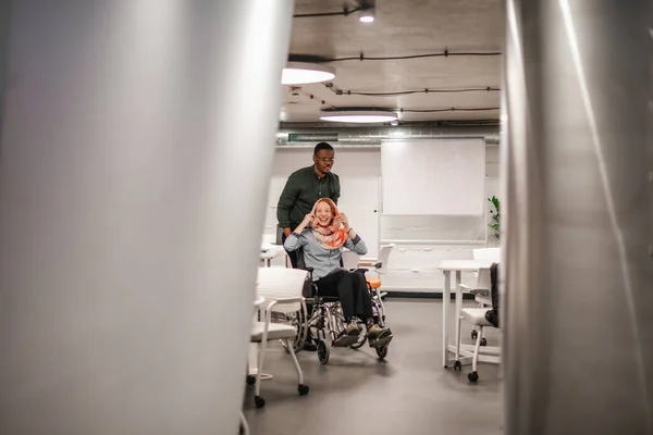 英俊的男性把他快乐的女同事推着轮椅穿过办公室 — 图库照片