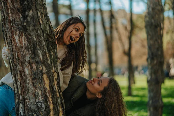 两个可爱的女孩在公园的树后玩耍和躲藏 卷发女孩一边唱歌一边望着他的朋友 — 图库照片