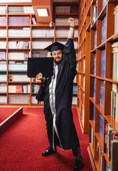 年轻而兴奋的毕业生一边拿着学士学位一边举手投足 头戴帽子 身穿长袍 — 图库照片