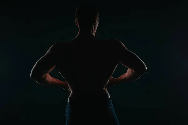 Well-built and charming man standing in front of a dark background and flexing his back