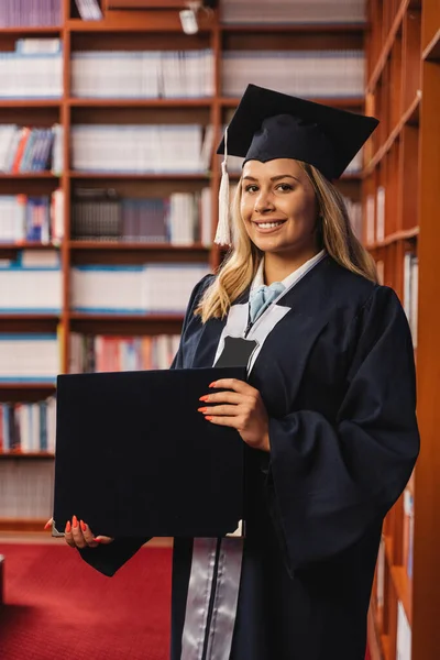 若くて幸せな卒業生はキャップとガウンを身に着け 図書館の中に彼女の学士号を保持しています — ストック写真