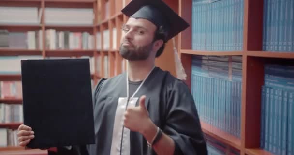 幸せな男性学生は 彼の卒業証書を保持しながら それを達成するために幸せな親指を示しています 大学図書館で彼の業績を披露する誇りある学生 — ストック動画