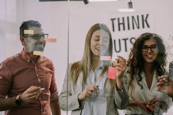 3人のビジネスマン 2人の女性1人の男性 笑顔でアイデアやシンボルをガラスの壁に書いてオフィスでポストカード — ストック写真