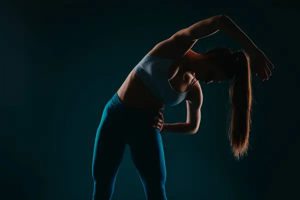 A confident, fit woman in a dark studio, doing exercises to transform her body. She focuses on her abs, biceps, chest, and triceps, showcasing her progress and inspiring others.