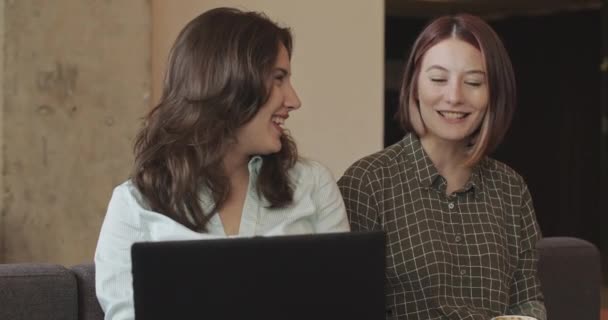 2人の女性の同僚がプロジェクトについて話し 快適なオフィスのソファー カメラのトラックの動きに休息しながら大声で微笑みます — ストック動画