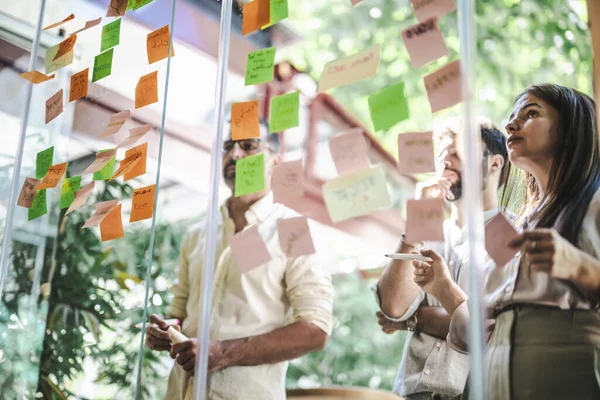 近代的なオフィスでの多種多様なビジネスチーム 戦略化 継続的にプロジェクトに取り組んで 粘着性のあるノートに覆われたガラス壁は 彼らの創造的なアイデアと進歩を反映しています — ストック写真
