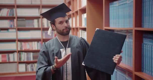 卒業した卒業証書を見せる幸せな男性学生は 達成するのが難しくないかのように見えます 大学図書館で彼の業績を披露する誇りある学生 — ストック動画