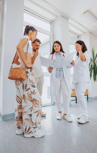 A group of doctors and specialists discussing diagnoses and treatment options with a happy couple in an indoor office setting. Medical results and expertise inform the conversation.