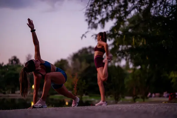 Fit women exercising in a green park at night, stretching and warming up together. Their athletic bodies showcase strength and flexibility as they prepare for a workout outdoors, near a small lake.