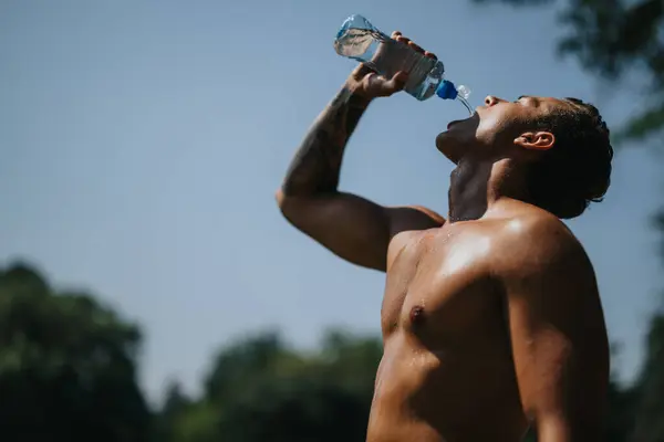 stock image A fit Caucasian athlete rests in a park, enjoying a sunny day. He drinks water, showcasing his strong muscles. Nature inspires his goal for a better body shape.