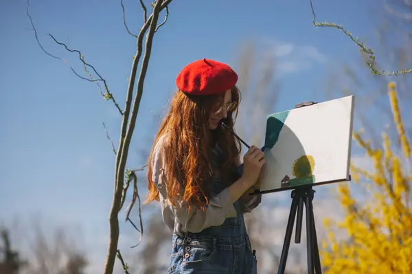 Red haired woman wearing red hat painting a colorful landscape while standing at the park