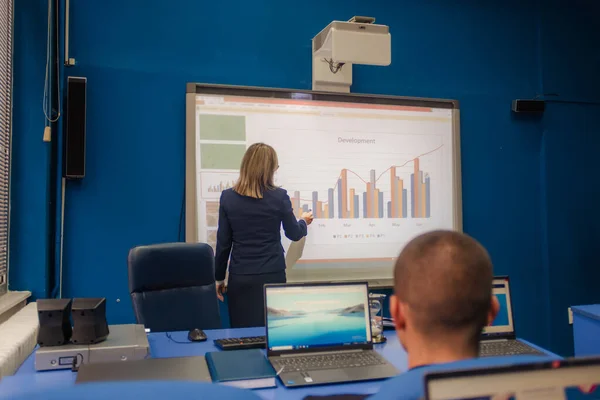Female college professor writing on smart board, delivering lecture to students, explaining math, economy, development, charts.