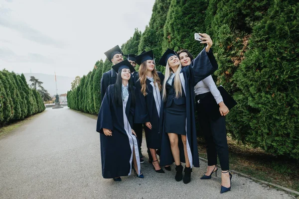 公園の卒業生は 友人と彼らのマイルストーンを祝い 彼らのガウンと帽子で卒業の思い出を作成します 卒業制服で一緒に立っている間 セルフィーを取る — ストック写真