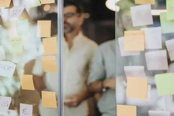 A diverse business team brainstorming and collaborating in a co-working space. Writing on sticky notes, they share ideas and discuss project costs and strategies on a glass wall.