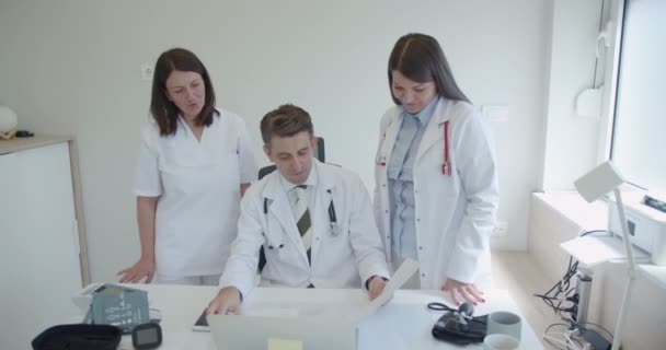 2人の医師が患者の結果について相談したり 経験豊富な男性医師に相談したりします 3人の医師が小さな近代的なオフィスで医学的結果を分析している — ストック動画