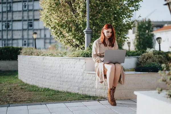 Redhead businesswoman working on lap top while sitting in urban city park on a beautiful sunny day.