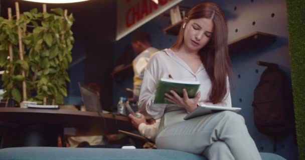 三十多岁的女商人坐在门边的沙发上 一边在笔记本上写下自己的商业想法 一边和队友分开工作 — 图库视频影像