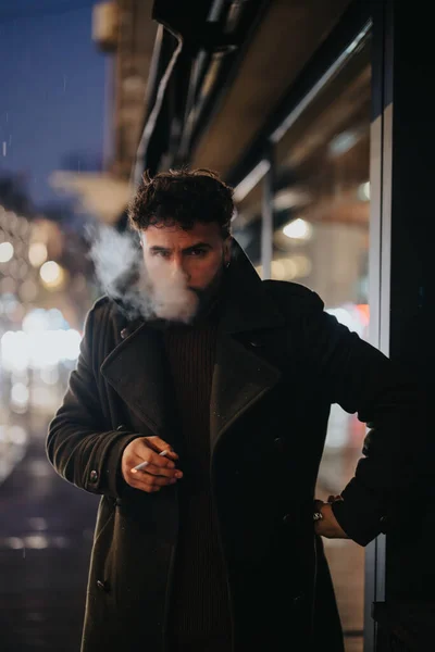 在一个繁华的城市里 一位时尚俊俏的商人站在市中心的人行道上 一边抽烟 一边在雨夜等出租车 — 图库照片