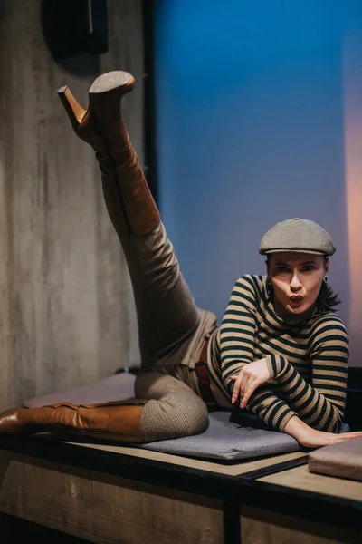 A stylish young woman in casual wear sits indoors, exuding beauty and charm as she poses with one leg up. With a trendy newsboy hat and a cheerful expression, she showcases her fashionable personality