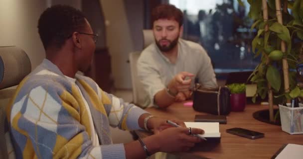 白人と黒人の2人の男性従業員が 現代のオフィスで一緒にテーブルに集まりました オフィスで一緒に働いている間 コーカサスの従業員は同僚と冗談を言った 彼らは笑って楽しむ — ストック動画