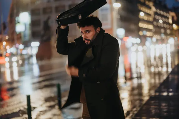 一个英俊的商人晚上站在一个繁忙的城市里 用手提电脑蒙着头 不让雨淋着 他正在等出租车下班回家 — 图库照片