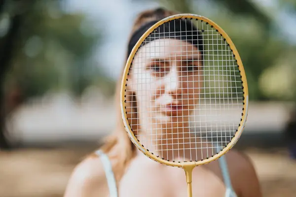 A photo of woman holding a racket with her face behind it. Badminton player posing with racket. Focus on racket.
