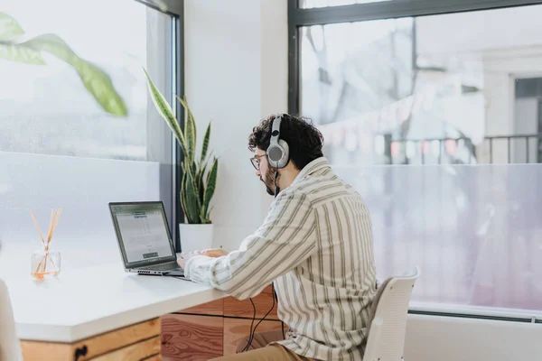 Male employee sitting on working desk next to the window is listening to music on the headphones and working on his daily tasks
