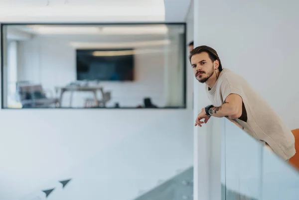 A thoughtful man with a beard leans on a glass partition in a modern office space, looking into the distance. Blurred office environment in the background evokes a sense of contemplation and focus.