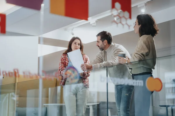Three professionals engaged in a conversation with reports in hand in a bright office corridor. The transparent glass partitions give a glimpse into the collaborative corporate environment.