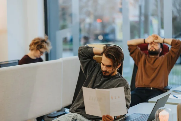 Three coworkers in an office experiencing different stages of stress and exhaustion, with one focused on work, another stretching, and the third deep in thought.