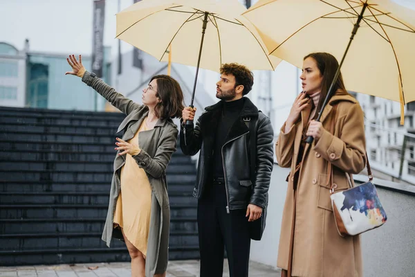 3人の友人が黄色い傘を持って 濡れた街路で待っている 彼らは たぶんタクシーやバスのために 異なる方向に期待して見えます — ストック写真
