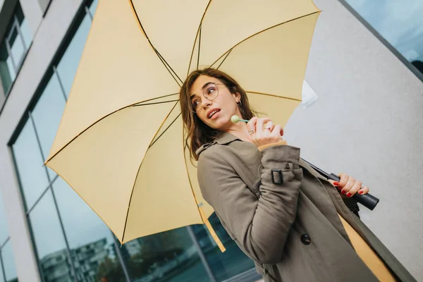 メガネをかけたスタイリッシュな若い女性が黄色い傘を持ち モダンな建築で都会的な背景に思慮深く見えます — ストック写真