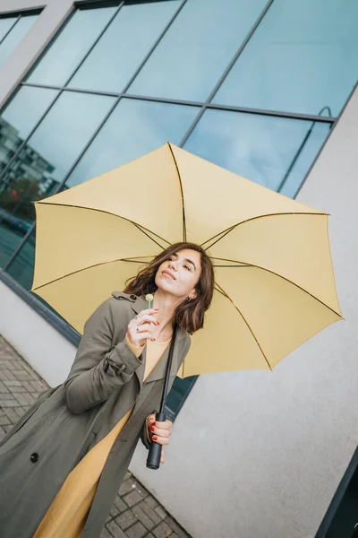 モダンなガラスの建物の背景に立ち 都会的なスタイルとエレガンスを例証する 黄色の傘を保持するファッショナブルな女性 — ストック写真