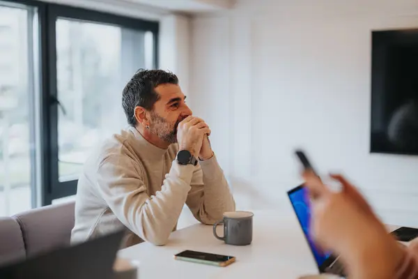カジュアルなセーターの高級男性は スマートフォンを使用して同僚との会議中に熟考しながら 明るいオフィスセッティングに座っています — ストック写真