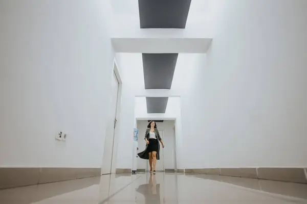 Elegant young woman in business attire walking with confidence down a bright, contemporary office hallway with a minimalist design.