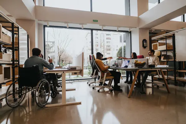 社員の多種多様なグループが 明るいオフィススペースでテーブルの周りに協力し 車椅子のアクセシビリティとチームワークを紹介しています — ストック写真