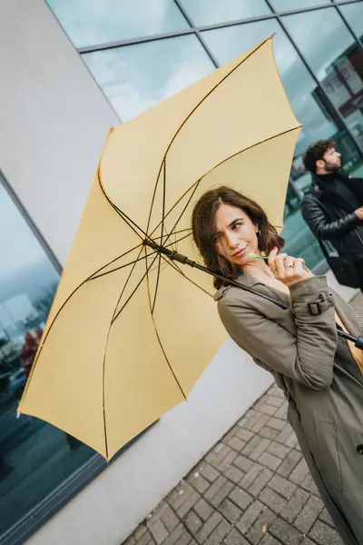 明るい黄色の傘を持っているファッショナブルな若い女性は カメラを眺めながらモダンな建物の前に立っています — ストック写真