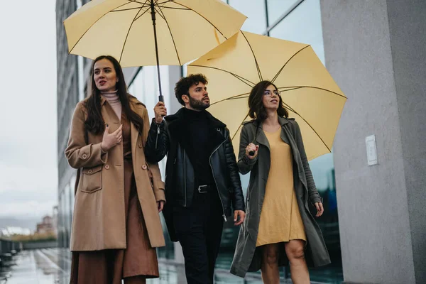3人のファッショナブルな友人は 明るい黄色の傘とスタイリッシュな衣装で武装した乾燥した都市のシーンをナビゲートし カジュアルな自信の空気を除きます — ストック写真