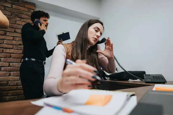 Busy office atmosphere with a focused woman multitasking on the phone and a man in the background on a call.