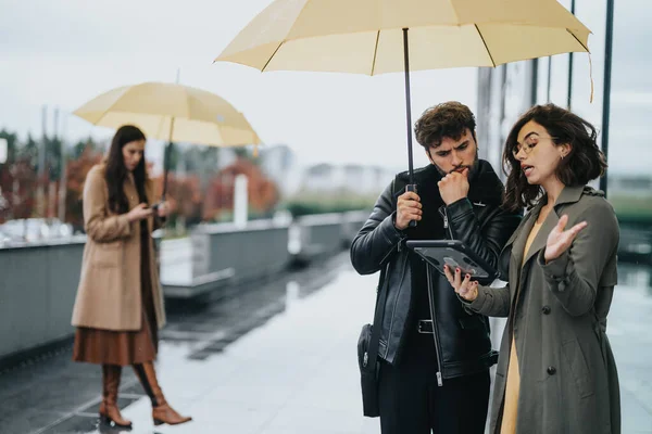 黄色い傘の下の2人は 雨を無視してタブレットで深刻なビジネスディスカッションに囲まれました — ストック写真